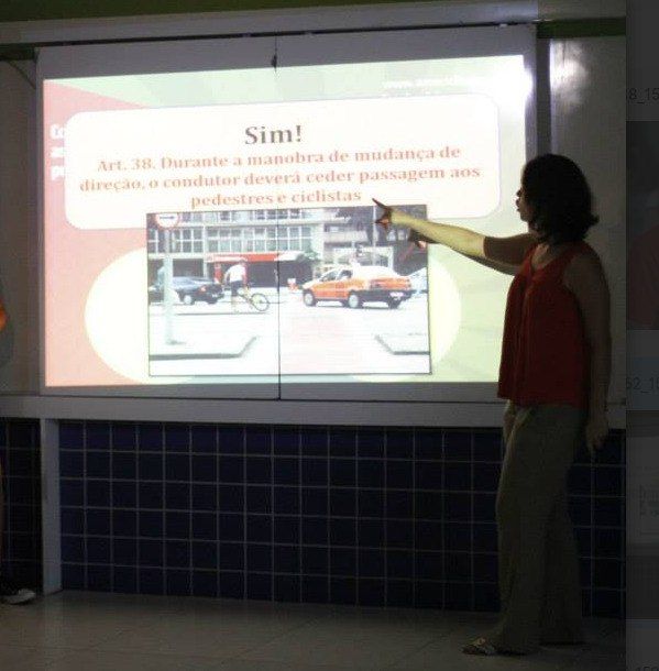 Pessoa apresenta atividade educativa da Ameciclo em sala de aula, com projeção de slides sobre mobilidade por bicicleta